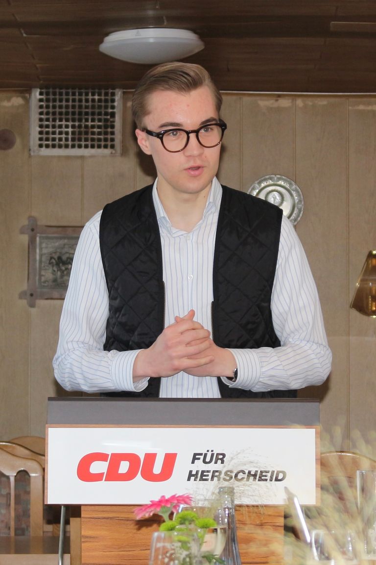 Person mit Brille in CDU-Vest vor Podium mit „CDU für Herscheid“-Schild spricht.